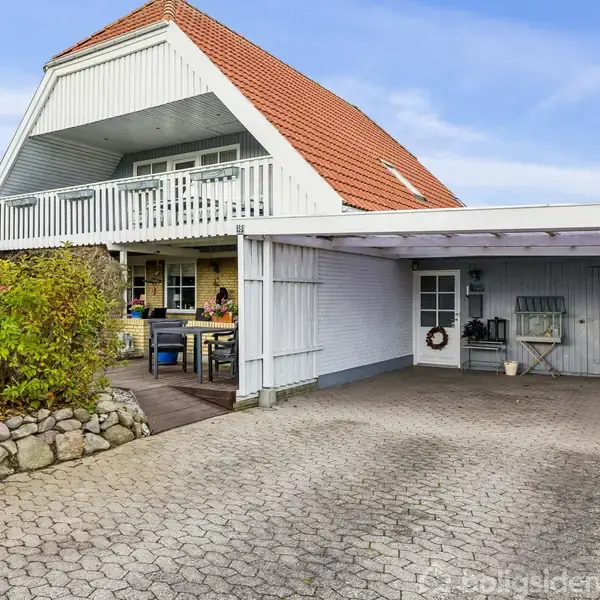 Hus med rød tag og hvid altan står foran en carport. Ved siden af er en terrasse med bord og stole. Hækken omkranser grunden og skaber en hyggelig atmosfære.