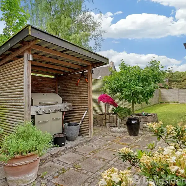 Grill under halvtag står på flisebelagt terrasse, omgivet af blomster og grønne planter i krukker, mens en stor have med træer og buske strækker sig i baggrunden under blå himmel med skyer.