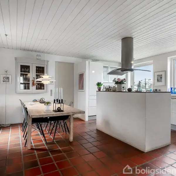 Spisebord med stole står i et lyst køkken-alrum med terracotta-fliser på gulvet. Rummet har hvide vægge og loft, et køkkenområde med en emhætte og store vinduer uden gardiner.