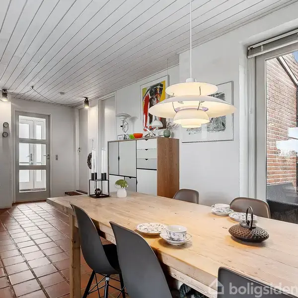En stor spisebord med seks stole er midtpunktet i et rummeligt rum. Loftslampen hænger over bordet. I baggrunden ses en kommode og kunst på væggen.