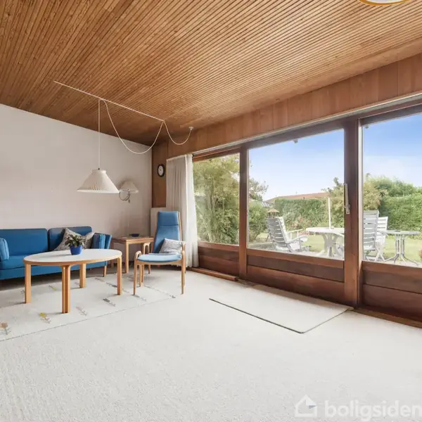Stue med blå sofa og stol ved siden af et træbord, vendt mod store vinduer med udsigt til en terrasse og have. Loftet har træpaneler, gulvet er lyst.