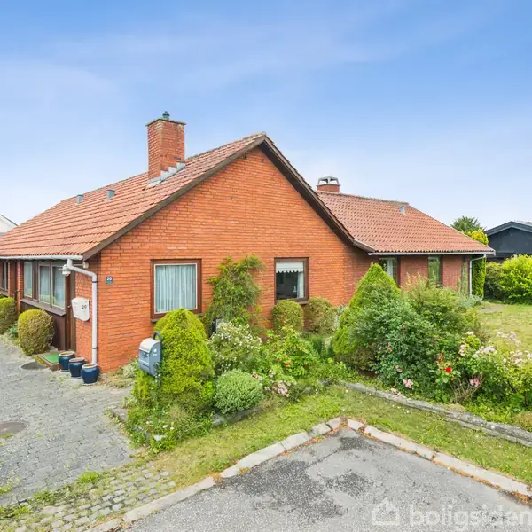 Rødt murstenshus med rødt tegltag, omgivet af velplejede buske og frodig have. Indkørsel af brosten fører til carport ved siden af huset. Klar blå himmel.