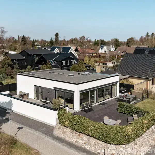 Fladt taghus med store vinduer står på en velholdt grund, omkranset af hække. Beliggende i et parcelhuskvarter med træer og nabohuse, er der en parkeret bil ved siden af huset.