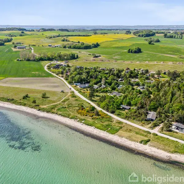 Kystlinje med grusstrand grænser til klart, lyseblåt havvand. Bebyggelse og grøn skov løber langs en landsvej. Landbrugsmarker strækker sig mod horisonten under en blå himmel.