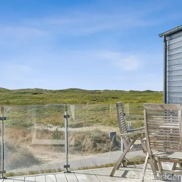 Terrassemøbler af træ står på en altan med glasværn. De er rettet mod et åbent landskab med grønne bakker under en klar blå himmel.