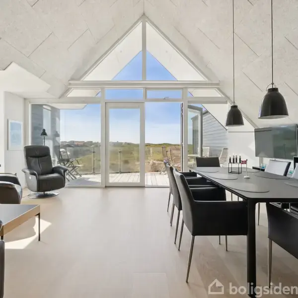 Stue med store vinduer og skrå loft, der lader lys strømme ind. Spisebord med sorte stole, to lænestole, og udsigt til terrasse og natur udenfor.