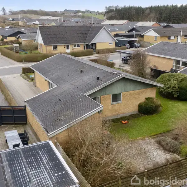 Et parcelhus med gråt tag er centralt placeret i en villakvarter. Huset er omgivet af grønne buske og græsplæne, med en flisebelagt terrasse og indkørsel i nærheden.
