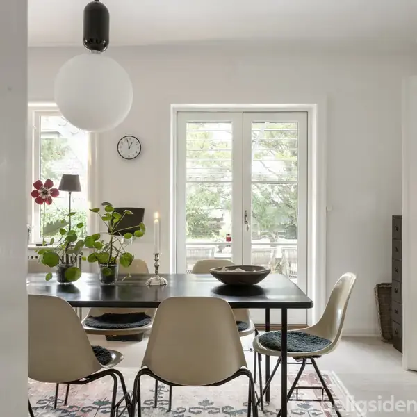 Et spisebord med stole er centralt placeret i et lyst rum. En stor lampe hænger over bordet, og der er planter og dekorationer. En dør åbner ud til en terrasse.