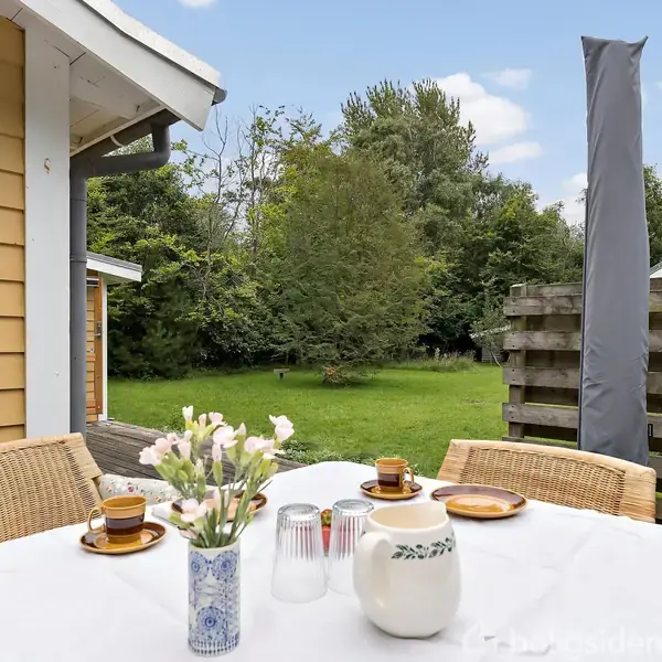 Bordet er dækket med en hvid dug, keramikkande, kopper og tallerkener. Kurvestole omgiver bordet. I baggrunden ses en græsplæne med træer og en delvist skjult bygning.
