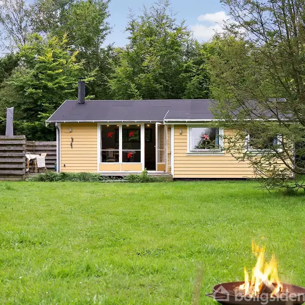 Et gult sommerhus står stille i en grøn have omgivet af træer. I forgrunden ses en ild i en skål.