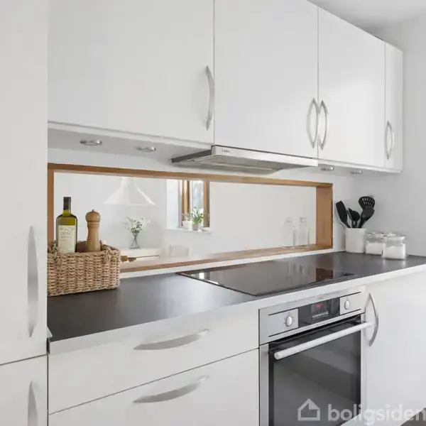 Hvide køkkenskabe omgiver en moderne indbygningsovn med en sort kogeplade. En kurv med olier står ved siden af kogepladen, og der er en trærammet spejlbagvæg. Et vindue til højre giver udsigt til natur.