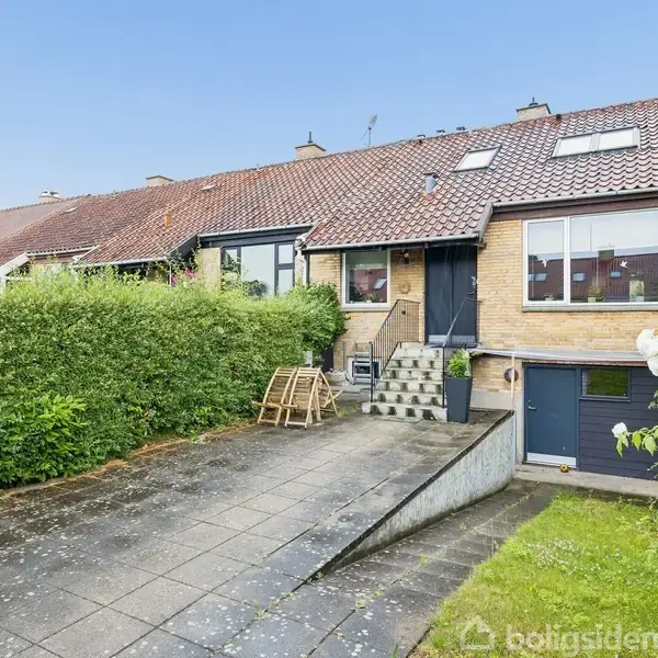 Rækkehus i mursten med rødt tegltag, placeret i række af lignende huse. Der er en indkørsel foran, omkranset af hæk og plæne med blomster. Træstol på terrasse.