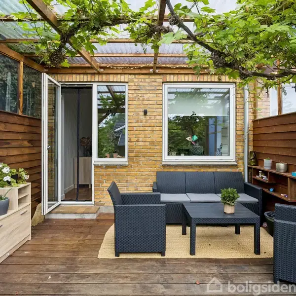 En terrasse med sorte havemøbler bestående af sofa, stole og bord under en pergola dækket af grønne planter. Omgivelserne har brune trævægge og et gulv af træplanker.