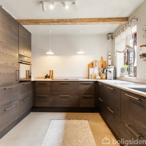 Moderne køkken med mørke træskabe og indbyggede apparater, oplyst af hængelamper, med træbjælker i loftet og vinduer med gardiner, der skaber en lys og indbydende atmosfære.