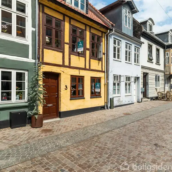 Gult bindingsværkshus med brune døre og vinduer. Der hænger et skilt med teksten “TIL SALG” på vinduet. Huset står på en brostensbelagt gade flankeret af andre farverige huse.