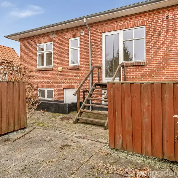 Rødt murstenshus med vinduer og lille trappe, der fører til en terrasse. Omkringliggende hegn og flisebelagt gårdhave med sporadisk beplantning. Klar himmel i baggrunden.