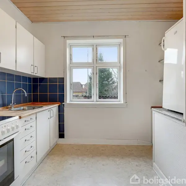 Et lyst køkken med hvide skabe og en blå flisevæg. En komfur og vask er placeret langs væggen. Et vindue viser udsigt over et træ og himmel.