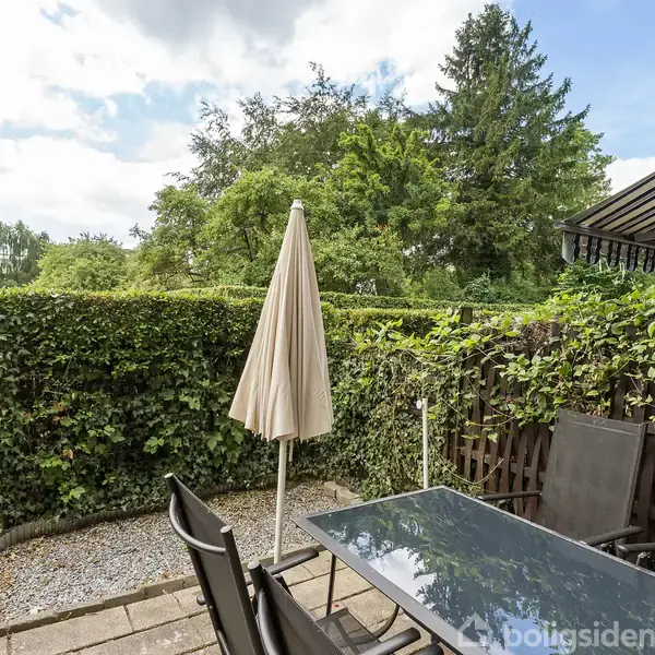 Terrassemøbler står omkring et glasbord under en lukket parasol på en terrasse. Terrassen er omgivet af en hæk og en høj trævæg, med udsigt til træer i baggrunden.
