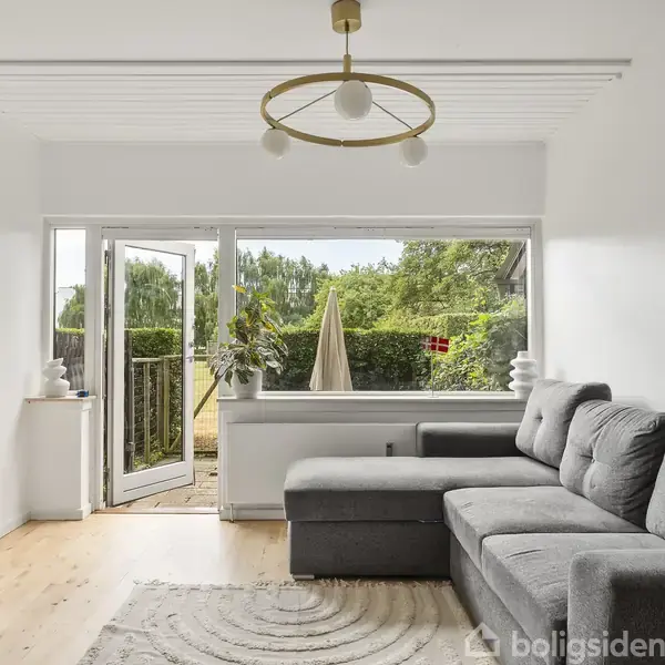 Grå sofa står i en stue med lyse trægulve, ved siden af et åbent vindue, der fører ud til en terrasse med grøn bevoksning udenfor.