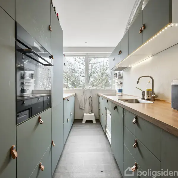 Et moderne køkken med grønne skabe, og en træbordplade. Ovn og mikroovn er indbygget i skabene. Store vinduer i baggrunden giver udsigt til træer.