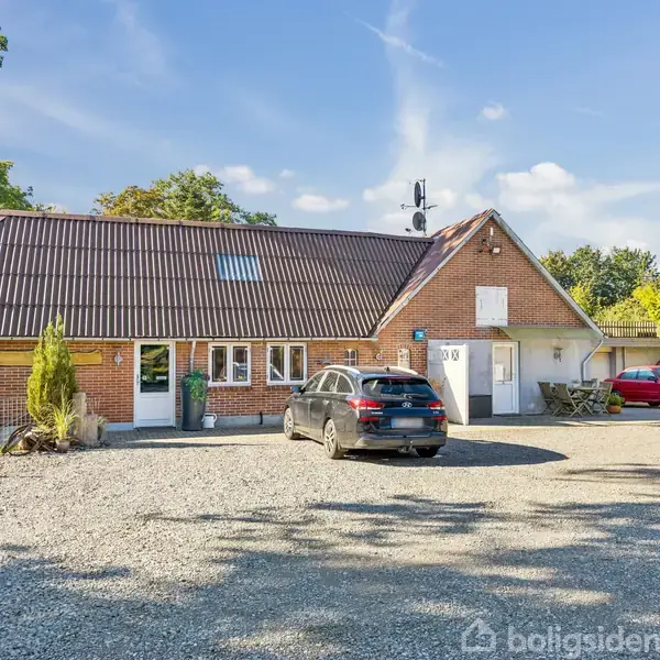 En bil holder parkeret foran et rødt murstenshus med skråt tag, omgivet af grus og træer i baggrunden under en klar blå himmel.