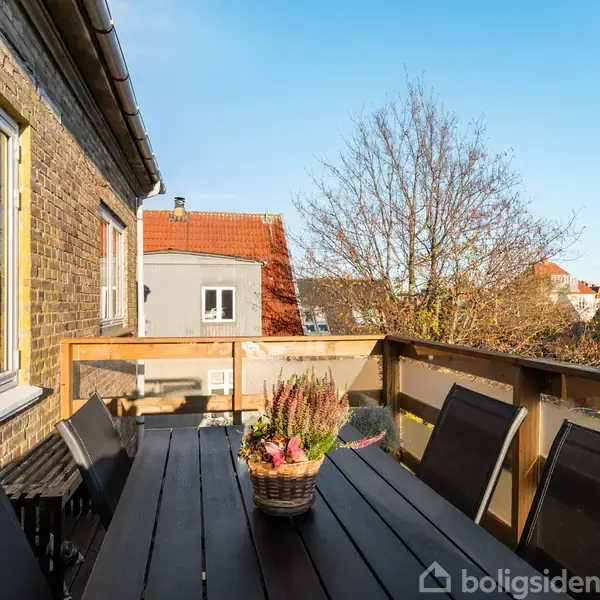 Et bord med sorte stole står på en altan. En blomsterkurv pynter bordet. I baggrunden ses bygninger og træer under en blå himmel.