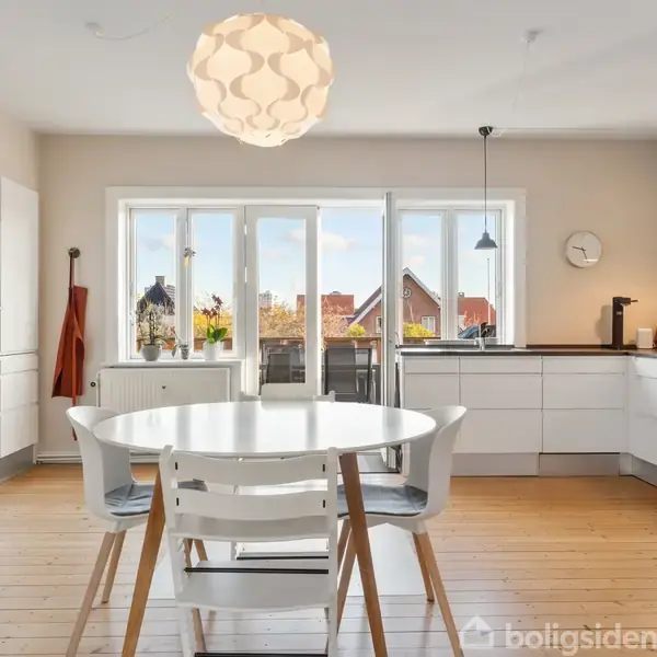 Et lyst køkken med et rundt hvidt bord og fire stole står på et trægulv. Store vinduer viser huse udenfor. Moderne indretning med hvide skabe og hængende lamper.