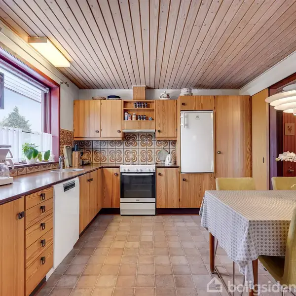 Et køkken med træskabe og fliser med mønster. Et bord med blomster og stole står i midten. Et komfur, køleskab og opvaskemaskine er placeret omkring rummet.