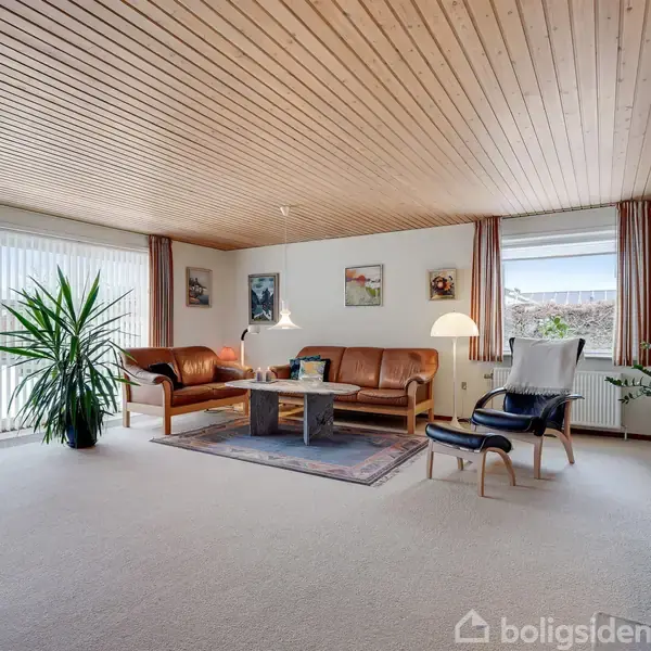 En stue med lædersofa og stol omkring et lavt bord på et tæppe, omgivet af planter og dekoreret med vægkunst. Store vinduer giver udsigt til det udendørs område.