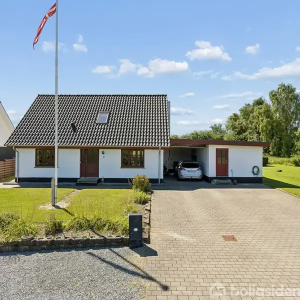 Et hvidt hus med gråt tag står stille på en velplejet græsplæne. En flagstang foran bærer et dansk flag. En indkørsel fører til en carport med en hvid bil.