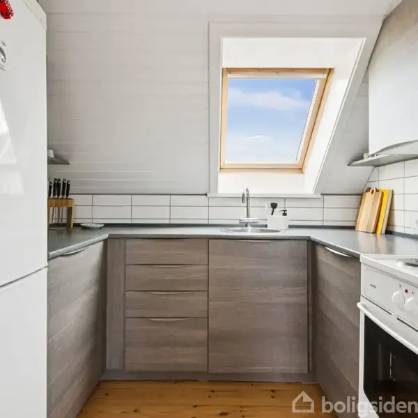 Et moderne køkken med grå skabe og træskuffer, hvor en hvid køleskab står til venstre. Der er et vindue ovenover vasken, og en ovn i hjørnet. Atmosfæren er lys og rummelig.