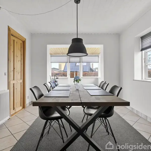 Et stort spisebord står i et lyst rum med seks stole rundt om. Bordet er dækket med bordskånere og en vase med blomster. Store vinduer og en radiator ses i baggrunden.