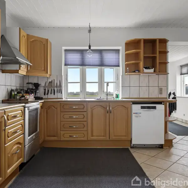 Køkken med træskabe og stålkomfur ligger i et solbelyst rum. En opvaskemaskine står ved et vindue med udsigt til natur. Fliser og måtte dækker gulvet.