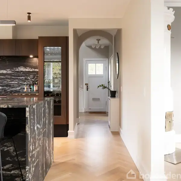 Køkken med marmor-bar, sorte stole og mørke trækabinetter. I baggrunden ses en gang med buet åbning og dør. Lys gulvbelægning og sofahjørne til højre.