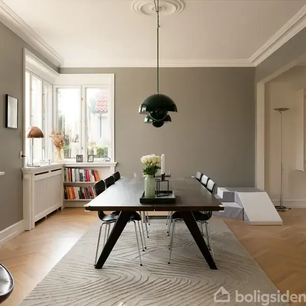 Et spisebord med en grøn pendellampe hænger over det i et stilfuldt, moderne rum. Bøger og planter er placeret ved vinduet. Grå vægge og et mønstret tæppe fuldender rummet.