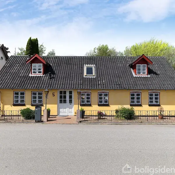 Et gulmalet hus med sorte tegl på taget står ved en stille vej. Huset har røde kvistvinduer og nummeret "5" ved hoveddøren, omgivet af små buske og blomsterbede.