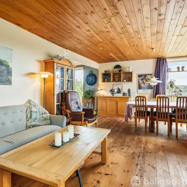 En stue med trægulv og træloft; en grå sofa og sofabord med stearinlys foran. I baggrunden står et spisebord med seks stole ved et vindue med gardiner.