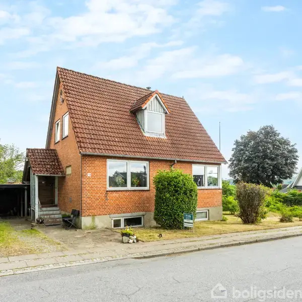 En rød murstenshus med en rødbrun tegltag. Husets forside vender mod en vej med fortov. Til venstre er en indkørsel med overdækket carport. Buske og træer omgiver huset.