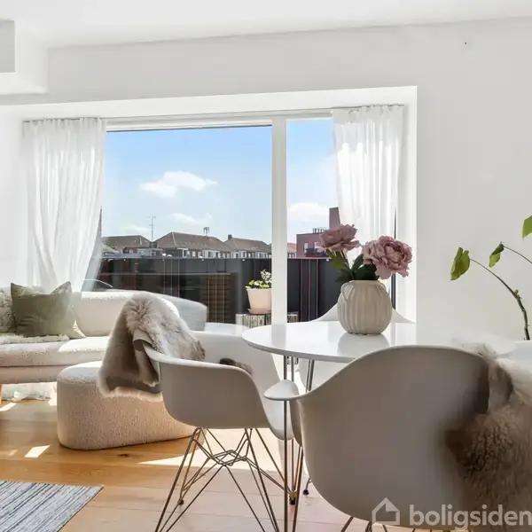 Stue med moderne møbler arrangeret omkring et rundt spisebord. Store vinduer lader sollys strømme ind. Blomster og planter dekorerer rummet, der har udsigt til en terrasse og himmel.