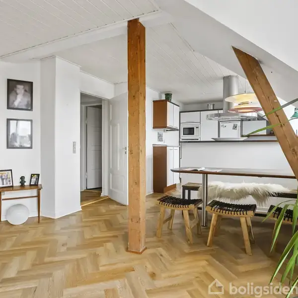 Et lyst køkkenalrum med trægulv, hvor et spisebord er flankeret af små bænke. En stor plante står ved vinduet, nær en åben reol og en moderne hvid gulvlampe.