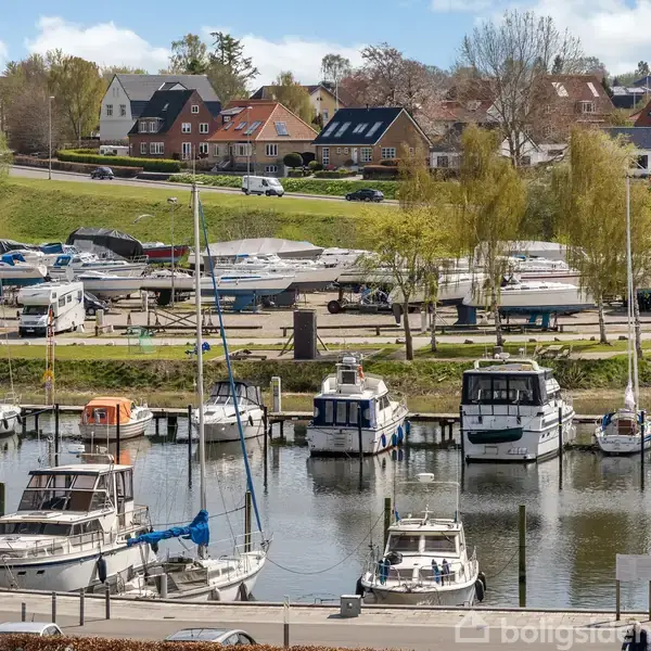 Både ligger forankret i en havn omgivet af grønne områder og huse i baggrunden. Træer og parkerede biler ses langs kajen.