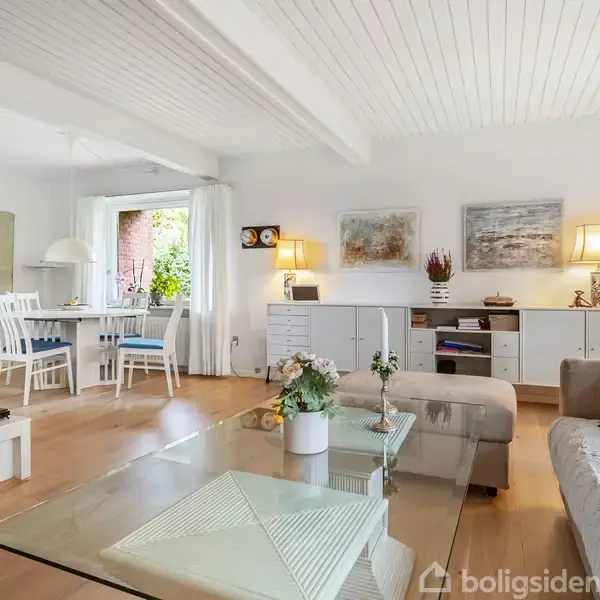 Stue med sofaer foran et sofabord, dekoreret med en vase. Til venstre er en spisestue med bord og stole. Rummet er lyst med hvide vægge og træloft.