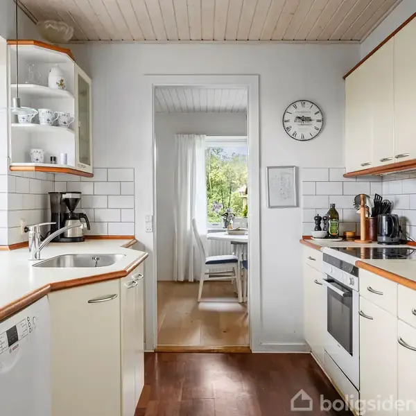 Et lyst køkken med hvide skabe og en indbygget ovn. En kaffemaskine står på bordet ved siden af vasken. Køkkenet åbner ind til en spiseplads med udsigt til grøn natur. En ur hænger på væggen.