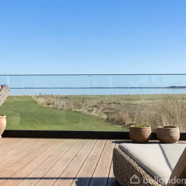 Terrassemøbler står stille på en træterrasse. I baggrunden ses en græsplæne og en glasafskærmning med udsigt over et åbent landskab og vand i horisonten.