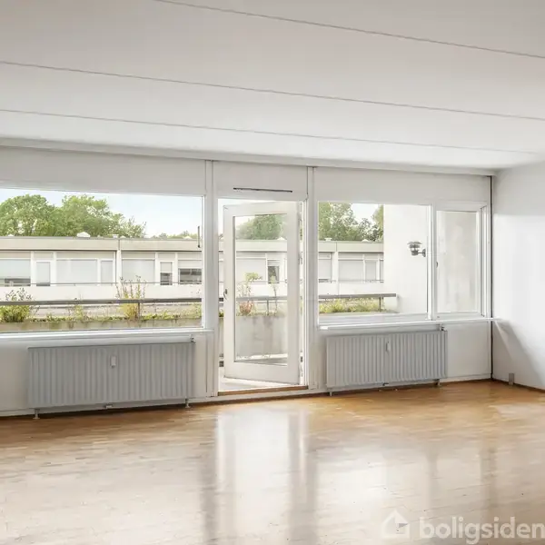 Et tomt værelse med trægulv og store vinduer fylder rummet med lys. Udenfor ses en bygning og grønne træer, hvilket giver en rolig atmosfære.