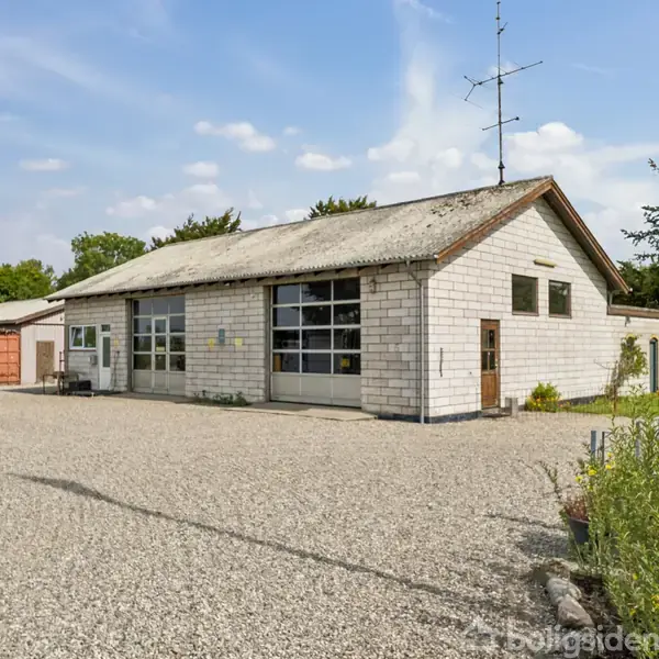 Gråt industribygning står stille i en grusbelagt gård med optrukne døre og træer i baggrunden under en klar blå himmel.