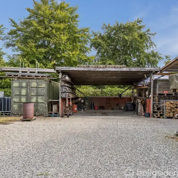 En åben carport med grusbelagt grund står ubenyttet midt i et gårdmiljø. Omgivet af forskellige opbevaringsmaterialer og træer, skaber det et rustikt og praktisk udseende.