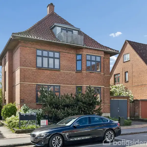 Rødt murstenshus med skråt tag står ved en vej. En mørk bil er parkeret foran huset, der er omgivet af buske og træer. En nabobygning ses i baggrunden.