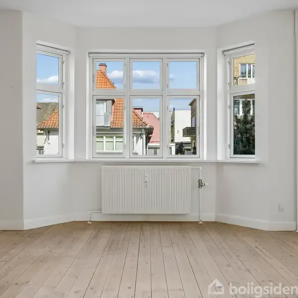 Et tomt rum med lyse træplankegulve og hvide vægge. Der er et stort vindue til højre, og en åben dør fører til et solbeskinnet rum med grønt udenfor.