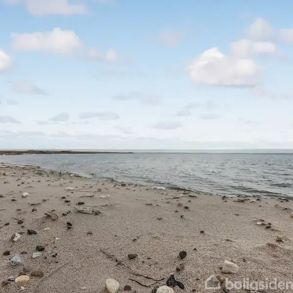 Strand med sand og spredte sten som fører til vandet. Havet ligger roligt under en blå himmel med spredte skyer. Kystlinjen strækker sig i det fjerne.